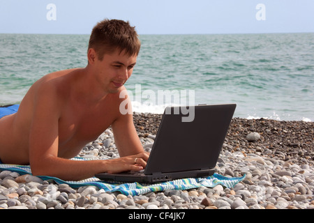 Jeune homme travaille sur son ordinateur portable sur littoral. portrait de guy avec laptop lying on beach. Banque D'Images