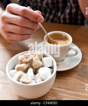 Détail de la main de l'homme tenant une cuillère en remuant une tasse de café expresso, avec un bol de sucre et des morceaux de sucre en premier plan. Banque D'Images