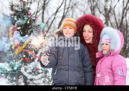 Mère de ses deux enfants est debout près de l'arbre de Noël à la lumière au Bengale en main en cloche hors focus. Banque D'Images