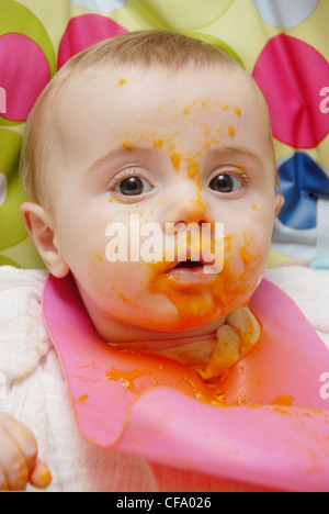 Un bébé de sexe féminin, les cheveux juste assis dans une chaise haute, le port d'un carré de mousseline rose et bib en plastique, manger en purée de couleur orange Banque D'Images