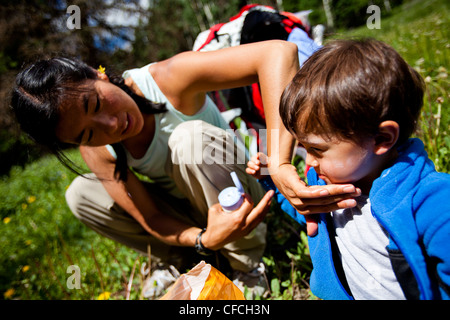 Une mère s'applique de la crème solaire à son fils, assis dans un pré herbeux, avec leur sac à dos derrière eux, sur la Williams Creek Tr Banque D'Images