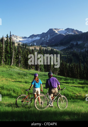 Jeune couple reste sur des vélos dans un pré, pic de Whistler, Colombie-Britannique, Canada. Banque D'Images