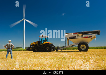 Un homme en champ d'orge récoltés avec le tracteur et le wagon de grain, l'affichage des éoliennes, près de St., Manitoba, Canada Banque D'Images