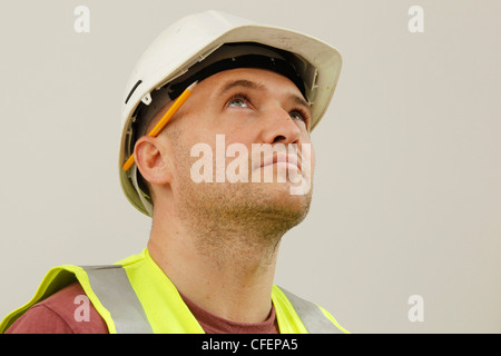 Un ouvrier pose modèle wearing hard-hat, hi-vis jacket et crayon derrière l'oreille Banque D'Images