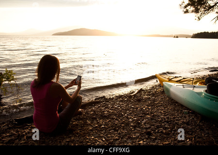 Une belle jeune femme jouant sur son téléphone cellulaire intelligent au coucher du soleil à côté d'un lac dans l'Idaho. Banque D'Images