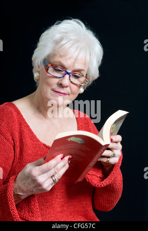 Attractive senior dame en vêtements lumineux assis sur une chaise reading book prises sur un fond noir Banque D'Images