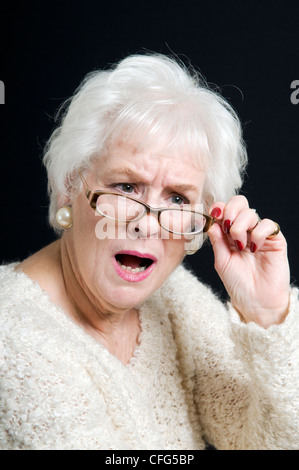 Senior lady avec un regard surpris sur le visage peering over verres pris sur un fond noir Banque D'Images