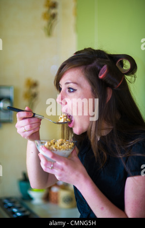 Jeune femme dans la vingtaine 20s ans wearing hair rollers manger le petit déjeuner en hâte pour se rendre au travail UK Banque D'Images