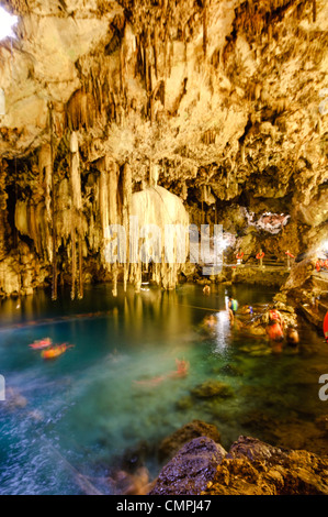 VALLADOLID, Mexique — les nageurs apprécient les eaux cristallines du Cenote X'kakah près de Valladolid, qui fait partie du vaste réseau de voies navigables souterraines du Yucatan. Le cenote, connu pour sa petite ouverture de plafond qui laisse entrer la lumière et la pluie, a de superbes stalactites suspendues à son plafond. Les nageurs, brouillés par une longue exposition, portent des gilets de sauvetage orange vif lorsqu'ils nagent dans l'eau constante à 76 degrés. Banque D'Images