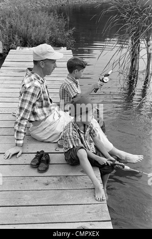 Années 1950 Années 1960 PÈRE AVEC DEUX FILS ASSIS SUR UN QUAI DE PÊCHE ENSEMBLE PISCINE PAR POND Banque D'Images