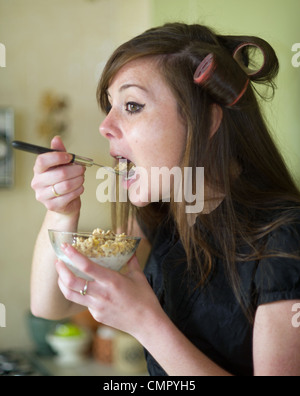 Jeune femme dans la vingtaine 20s ans wearing hair rollers manger le petit déjeuner en hâte pour se rendre au travail UK Banque D'Images