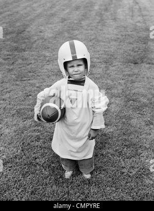 1950 BOY STANDING IN GRASS WEARING OVERSIZED SHIRT & HELMET HOLDING FOOTBALL Banque D'Images