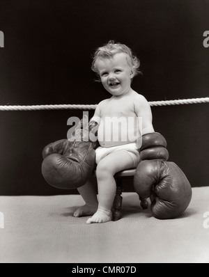 1930 SMILING BABY ASSIS SUR LA SELLE EN ring de boxe portant des gants de boxe surdimensionnés portant l'érythème Banque D'Images