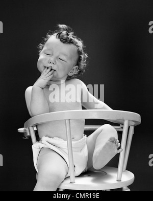 Années 1940 Années 1950 BABY SITTING EN ARRIÈRE dans une chaise haute avec une jambe dans l'air les yeux fermés et LA MAIN SUR LA BOUCHE Banque D'Images