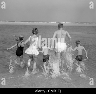 1960 VUE ARRIÈRE DE FAMILLE DE 5 HOLDING HANDS RUNNING INTO OCEAN Banque D'Images