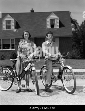 1950 SMILING TEENS SUR RUE DE BANLIEUE GARÇON ASSIS SUR LE VÉLO fille debout à côté de vélo avec panier Banque D'Images