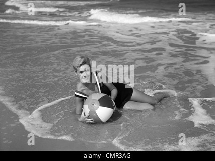 1960 FEMME EN MAILLOT DE COUCHÉ DANS LE SURF tenant un ballon de plage EN PLEIN AIR souriant Banque D'Images