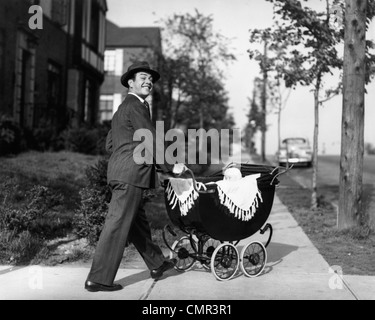 Années 1940 Années 1950 SMILING PÈRE EN FONCTION & HAT BÉBÉ EN POUSSANT VERS LE BAS DU CHARIOT TROTTOIR LOOKING AT CAMERA Banque D'Images