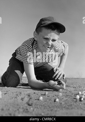 Années 1950 Années 1960 Garçon jouant aux billes à genoux dans la terre de loucher manque une dent de devant Banque D'Images