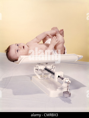 Années 1950 Années 1960 SMILING BABY LYING ON WEIGHT SCALE Banque D'Images