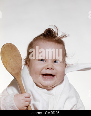 Années 1960 Années 1970 SMILING BABY WEARING BIB ET HOLDING cuillère en bois Banque D'Images