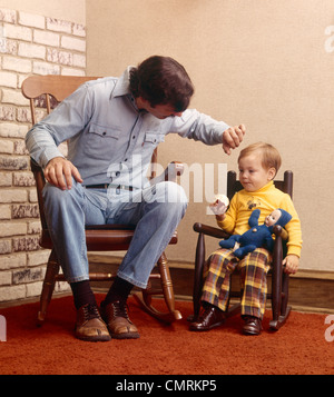 1970 1970 RETRO père assis dans un fauteuil à bascule à côté de son tout-petit à petit fauteuil à bascule poupée EATING APPLE Banque D'Images