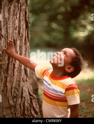 1970 1970 SMILING AFRICAN-AMERICAN BOY LOOKING UP TRONC DE L'ARBRE VERS LE HAUT À RAYURES LOOK TEE SHIRT RETRO EXTÉRIEUR Banque D'Images