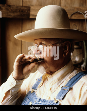 1960 SENIOR MAN WEARING HAT BORD LARGE LUNETTES RIM FIL BLEU JEAN À L'HARMONICA SÉANCE RÉVISIONS BIB Banque D'Images