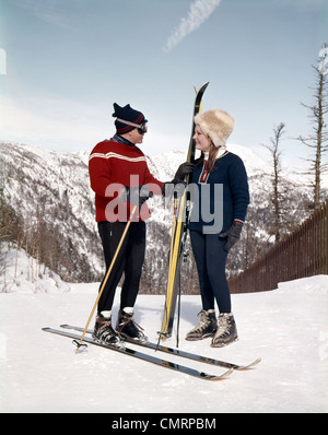 1960 1960 RETRO SMILING COUPLE HOMME FEMME EN CONVERSATION SUR LA PENTE DE SKI WEARING SKI GEAR HAT Gants Lunettes de ski montagne Banque D'Images