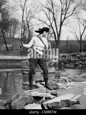 Années 1920 WOMAN IN HAT AVEC FILET DE PÊCHE & TACKLE BAG ATTACHÉ À SON CASTING HORS LIGNE DANS LE RUISSEAU Banque D'Images