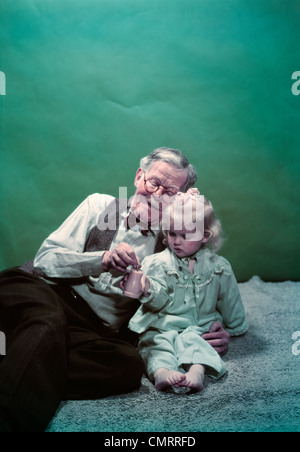 Années 1940 Années 1950 GRAND-PÈRE ASSIS SUR ÉTAGE AVEC PETITE-FILLE METTRE DES PIÈCES DANS LA BANQUE DE JOUETS Banque D'Images