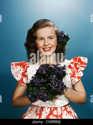 Années 1940 Années 1950 SMILING YOUNG WOMAN WEARING RED ET WHITE DRESS HOLDING bouquet de violettes Banque D'Images