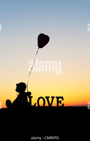 Ours en peluche tenant un ballon en forme de coeur trop suivant le mot amour au lever du soleil Banque D'Images