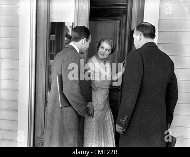 Années 1930 Années 1940 DEUX PORTE À PORTE VENDEURS PARLANT À LA FEMME AU FOYER À L'AVANT PORTE EN PRÉSENTATION DE VENTE Banque D'Images