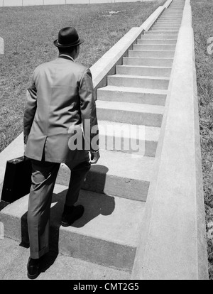1960 BUSINESS MAN IN SUIT HOLDING BRIEFCASE FEDORA & DÉMARRAGE LONG VOL D'ESCALIERS DE CIMENT DEVANT LUI. Banque D'Images
