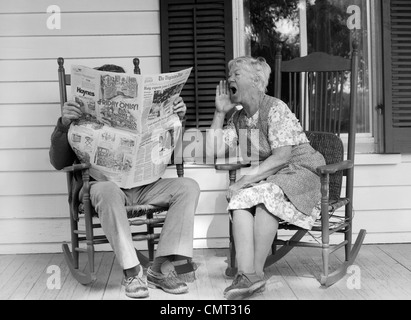 1970 COUPLE DE PERSONNES ÂGÉES DANS DES CHAISES À BASCULE SUR LE PORCHE MAN READING NEWSPAPER TANDIS QU'épouse se lamente sur lui Banque D'Images