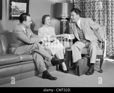 1950 COUPLE SITTING ON COUCH TALKING TO SMILING VENDEUR D'ASSURANCE DE LES REMETTRE UNE POLITIQUE Banque D'Images