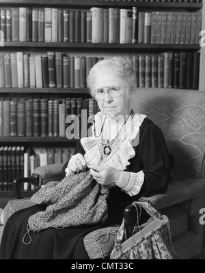 Années 1920 Années 1930 femme âgée assis devant BIBLIOTHÈQUE TRICOT EXPRESSION SÉRIEUSE LOOKING AT CAMERA Banque D'Images