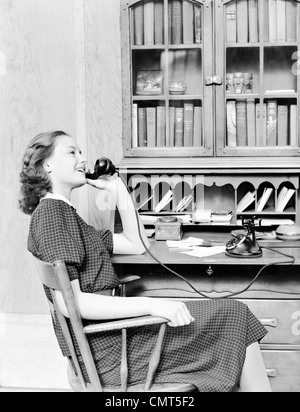 Années 1930 Années 1940 TEENAGE GIRL SITTING AT DESK TALKING ON vieux téléphone à cadran rotatif noir Banque D'Images