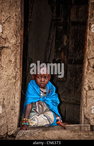 Enfant masaï, portant des vêtements traditionnels, assis dans l'embrasure d'une hutte de terre dans un village de la Masai Mara, Kenya, Afrique Banque D'Images