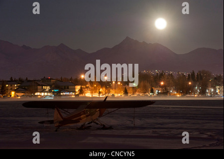 La pleine lune se lève sur les montagnes Chugach, avec un avion sur ski stationné sur le lac gelé du capot à l'avant-plan, l'Alaska, Winter Banque D'Images