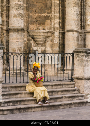 Un adulte femme cubaine habillée d'une robe jaune et jaune fantaisie foulard se trouve sur la cathédrale étapes avec des roses, La Havane, Cuba. Banque D'Images