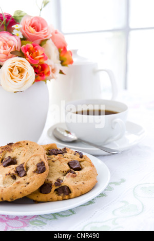 Biscuit aux brisures de chocolat sur plaque blanche avec du café noir et des fleurs Banque D'Images