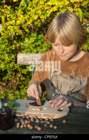 Le noisetier commun (Corylus avellana), garçon assis à la table de jardin Mise à tartiner au chocolat qu'il a faite de l'auto-prélèvement de noisettes, la poudre de cacao, de beurre et de sucre sur une tranche de pain, Allemagne Banque D'Images