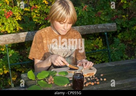 Le noisetier commun (Corylus avellana), garçon assis à la table de jardin Mise à tartiner au chocolat qu'il a faite de l'auto-prélèvement de noisettes, la poudre de cacao, de beurre et de sucre sur une tranche de pain, Allemagne Banque D'Images