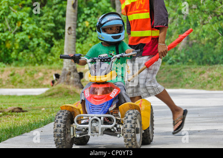 Petit garçon asiatique avec de grands enfants équitation casque quad ATV observé par commissaire de pistes avec drapeau rouge - Puerto Galera, Philippines Banque D'Images