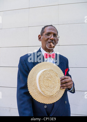 Un représentant de Cuba Personnes âgées sombre dans un chapeau canotier de paille, bleu sport manteau, noeud papillon rouge, et un cigare se distingue par un mur à La Havane. Banque D'Images