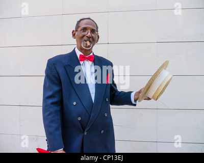 Un représentant de Cuba Personnes âgées sombre dans un chapeau canotier de paille, bleu sport manteau, noeud papillon rouge, et un cigare se distingue par un mur à La Havane. Banque D'Images