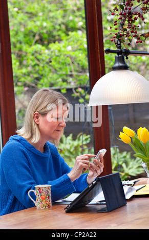 TABLETTE et SMARTPHONE WIFI HAUT DÉBIT femme blonde travaillant à la maison dans le jardin d'hiver en utilisant son Apple iPad Tablet PC et iPhone smartphone Banque D'Images