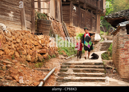 Une femme Miao basha transportant sur ses épaules avec des paniers de légumes et un enfant dans un village, Chine Banque D'Images